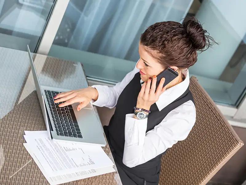 A woman in business attire uses a cellular phone while studying information on a laptop computer.