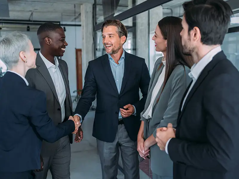 A group of five well-dressed business professionals shake hands while standing in a lobby.
