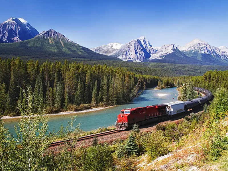 A freight train rounds the bend along a river in mountainous terrain.